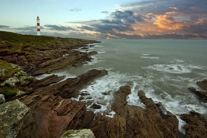 Leuchtturm auf einer felsigen Küste mit Blick auf ein aufgewühltes Meer bei Sonnenuntergang.