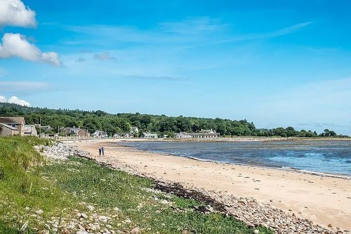 Küstenlandschaft mit Sandstrand, grüner Vegetation und fernen Gebäuden entlang der Küste.
